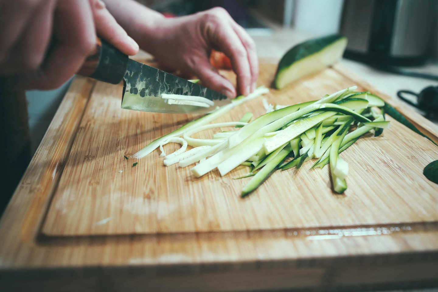 Hands slicing zucchini on a wooden cutting board.