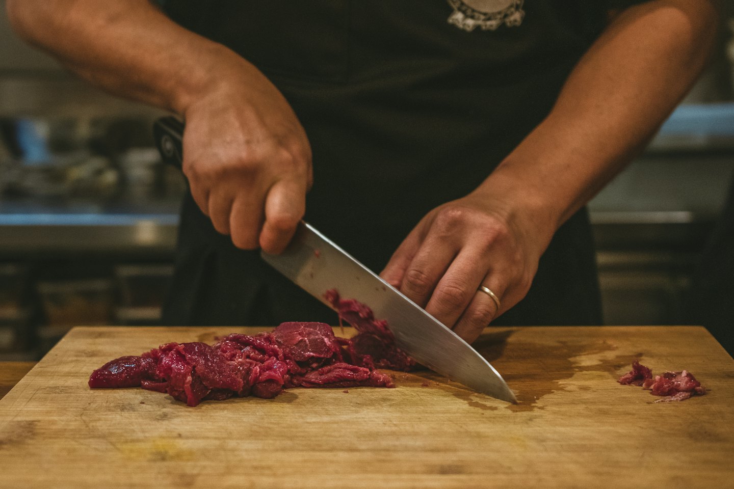 Hands in a dark apron chop raw meat on wood.